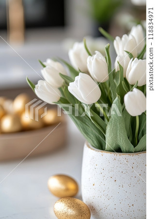 White tulips in a speckled vase beside golden eggs on a wooden platter in a modern kitchen 118834971