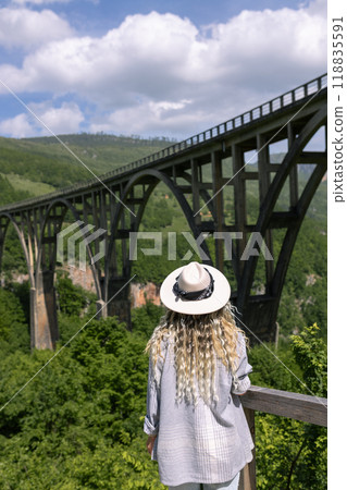 A girl in a hat stands against the backdrop of a beautiful arched bridge over the Tara River, a popular tourist attraction in Montenegro. A girl in a hat stands against the backdrop of a beautiful arched bridge over the Tara River, a popular tourist attraction in Montenegro. 118835591