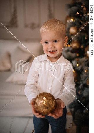 A young child holds a golden ornament while smiling in a cozy holiday setting at home 118836021