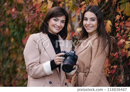 Girls are reviewing the photos they took on the camera. Two beautiful women stand in front of red and yellow trees.  118836487