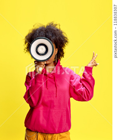 Stylish woman in pink hoodie holds megaphone, sending loud and confident message against vibrant studio background. Stylish woman in pink hoodie holds megaphone, sending loud and confident message against vibrant studio background. 118838307