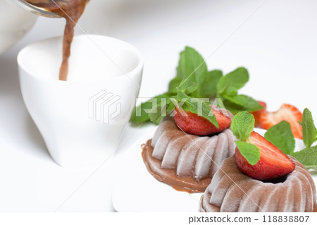 Chocolate fudge garnished with half a strawberry with petals. Garnished with sprigs of mint and strawberries. Nearby, they pour coffee into a cup. On the white surface of the table. 118838807