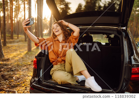 Happy stylish female tourist in trunk of a car with phone, taking a selfie, talking on a video call. 118839302