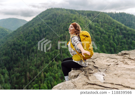 Beautiful traveler with a yellow hiking backpack observes the mountain scenery from a cliff. Beautiful traveler with a yellow hiking backpack observes the mountain scenery from a cliff. 118839566