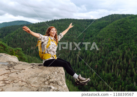 Beautiful traveler with a yellow hiking backpack observes the mountain scenery from a cliff. 118839567
