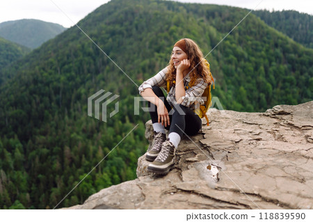 Beautiful traveler with a yellow hiking backpack observes the mountain scenery from a cliff. 118839590