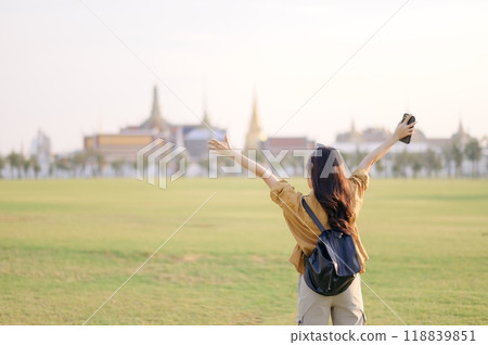 A Traveler Asian woman in 30s, bathed in golden glow of a Bangkok sunset, laughs with carefree joy, her arms outstretched as if to embrace freedom of the moment. Backpack strapped on. A Traveler Asian woman in 30s, bathed in golden glow of a Bangkok sunset, laughs with carefree joy, her arms outstretched as if to embrace freedom of the moment. Backpack strapped on. 118839851
