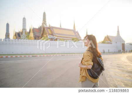 Traveler asian woman in her 30s, backpack slung over her shoulder, explores the intricate details of Wat Pra Kaew with childlike wonder. Sunlight dances on the golden rooftops. 118839863
