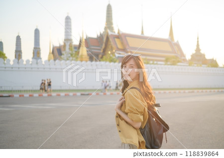 Traveler asian woman in her 30s, backpack slung over her shoulder, explores the intricate details of Wat Pra Kaew with childlike wonder. Sunlight dances on the golden rooftops. 118839864