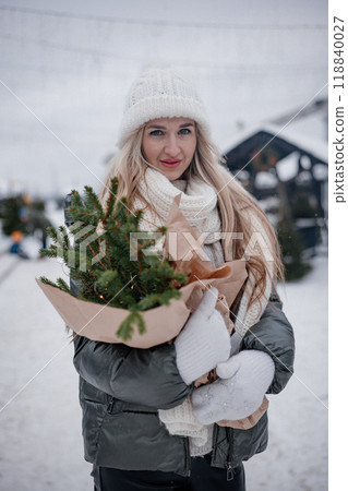 Portrait of young blond woman in beige coat walking in the city with a bouquet of fresh fir branches Portrait of young blond woman in beige coat walking in the city with a bouquet of fresh fir branches 118840027