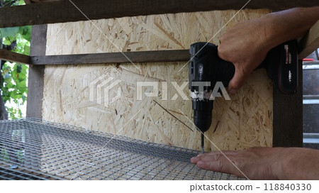 hands holding a screwdriver screwing fasteners into the structure of a cage for domestic quails or small animals, installing mesh into the structure of a cage for farm poultry hands holding a screwdriver screwing fasteners into the structure of a cage for domestic quails or small animals, installing mesh into the structure of a cage for farm poultry 118840330