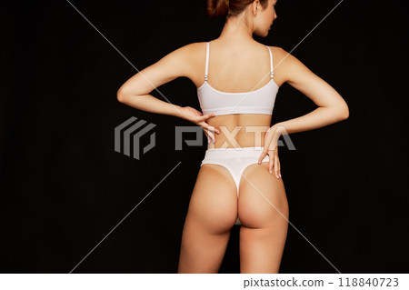 Rear view photo of young woman posing in white lingerie shoes her toned body curves against black studio background. 118840723