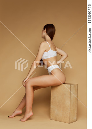 Female model poses seated on block, dressed underwear creating natural flow of lines against beige studio background. Female model poses seated on block, dressed underwear creating natural flow of lines against beige studio background. 118840726