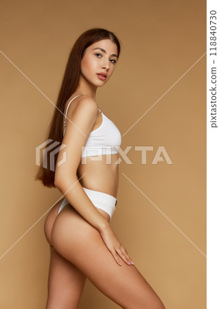 Side profile of young, slim attractive woman with long brunette hair posing in white underwear against beige studio background. 118840730