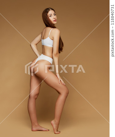 Full-length, side view photo of showcasing her sleek figure in white lingerie against beige studio background. Femininity. 118840731