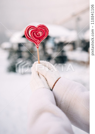 A person in white gloves holds a heart-shaped lollipop in a snowy winter setting 118841106