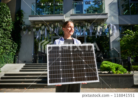 Woman holding solar panel for charging, stands outdoors, smiling in urban setting. Concept of green energy generation. Woman holding solar panel for charging, stands outdoors, smiling in urban setting. Concept of green energy generation. 118841551