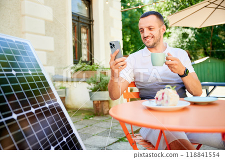 Young man charging smartphone from solar panel while sitting in cafe. Handsome male drinking coffee and checking his phone next to solar battery. Conscious male choosing sustainable lifestyle. Young man charging smartphone from solar panel while sitting in cafe. Handsome male drinking coffee and checking his phone next to solar battery. Conscious male choosing sustainable lifestyle. 118841554