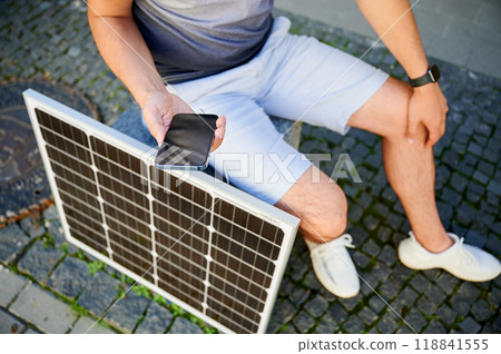 Cropped photo of man charging his smartphone from solar panel. Young man using portable solar battery to charge his device. Concept of alternative energy sources and sustainable lifestyle. Cropped photo of man charging his smartphone from solar panel. Young man using portable solar battery to charge his device. Concept of alternative energy sources and sustainable lifestyle. 118841555