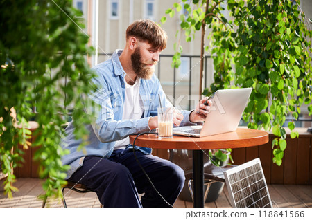 Man works on laptop and smartphone at outdoor table, with glass of iced coffee. Solar panel nearby, emphasizing sustainable, modern eco-friendly workspace that combines technology and sustainability. 118841566