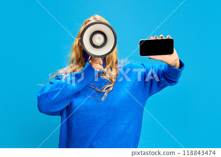 Young blonde woman in blue hoodie holding megaphone and mobile phone with blank screen against blue studio background. Online shopping Young blonde woman in blue hoodie holding megaphone and mobile phone with blank screen against blue studio background. Online shopping 118843474