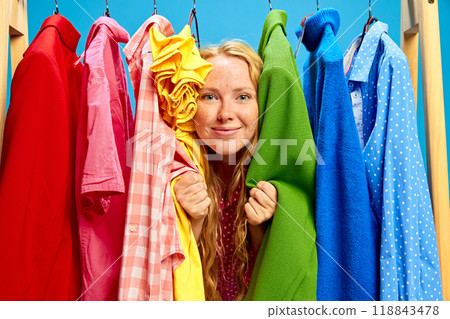 Cheerful and super excited young woman peeking through clothes rack against blue studio background. Limited-time offer ad Cheerful and super excited young woman peeking through clothes rack against blue studio background. Limited-time offer ad 118843478