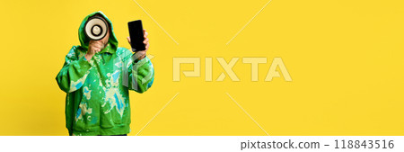 Man in green hoodie holding megaphone and showing mobile phone screen against yellow studio background. Limited-time offer promotion Man in green hoodie holding megaphone and showing mobile phone screen against yellow studio background. Limited-time offer promotion 118843516