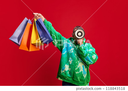Man in stylish hoodie holding colorful shopping bags and talking in megaphone against red studio background. Limited-Time Offers Man in stylish hoodie holding colorful shopping bags and talking in megaphone against red studio background. Limited-Time Offers 118843518