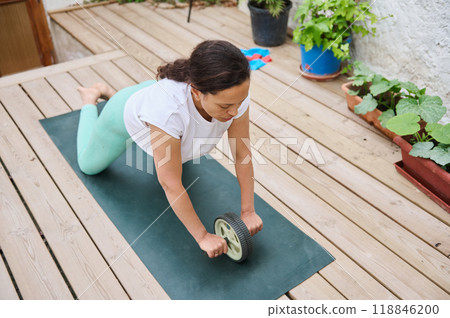Woman practicing yoga with ab roller on wooden deck, focusing on fitness and core strength outdoors in a peaceful garden setting Woman practicing yoga with ab roller on wooden deck, focusing on fitness and core strength outdoors in a peaceful garden setting 118846200