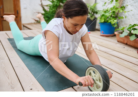 Woman practicing ab wheel workout on wooden deck in peaceful outdoor setting 118846201