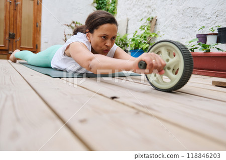 Determined woman exercising with an ab roller on a yoga mat in an outdoor setting, showing strength and concentration Determined woman exercising with an ab roller on a yoga mat in an outdoor setting, showing strength and concentration 118846203