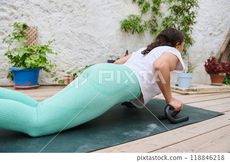 Woman exercising outdoors doing push-ups on a yoga mat in a garden setting with plants around, wearing green leggings for a healthy lifestyle concept. 118846218