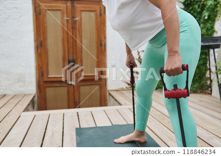 Woman exercising outdoors with resistance bands performing a bent-over row on a wooden deck in casual athletic wear. Woman exercising outdoors with resistance bands performing a bent-over row on a wooden deck in casual athletic wear. 118846234