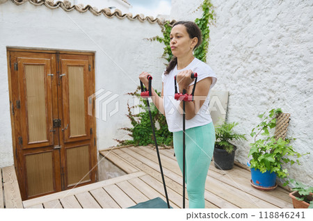 Woman exercising with resistance bands in a serene outdoor setting on a wooden deck with plants, focused on fitness and wellness. 118846241