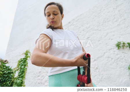 Woman exercising outdoors with resistance bands in a bright white and green environment, focused on her fitness routine 118846250