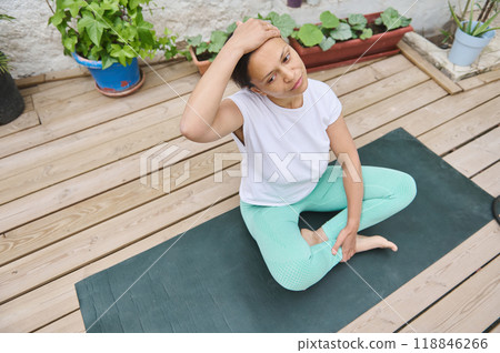 Woman practicing yoga on a wooden deck doing neck stretching exercises in a peaceful outdoor garden setting Woman practicing yoga on a wooden deck doing neck stretching exercises in a peaceful outdoor garden setting 118846266