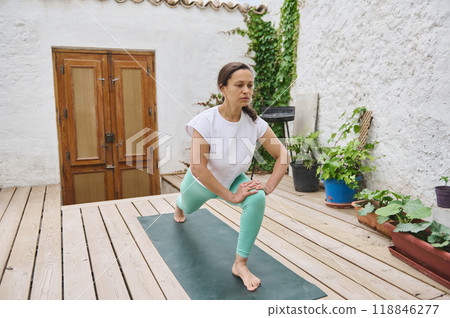 Woman practicing yoga in a serene outdoor space with plants, focusing on a deep lunge stretch on a wooden deck 118846277