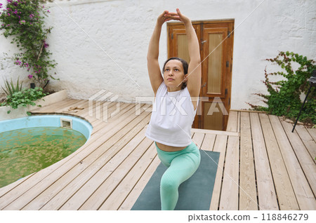 Woman practicing yoga outdoors performing a stretch on a wooden deck in a serene garden setting, focusing on wellness and mindfulness for a healthy lifestyle Woman practicing yoga outdoors performing a stretch on a wooden deck in a serene garden setting, focusing on wellness and mindfulness for a healthy lifestyle 118846279