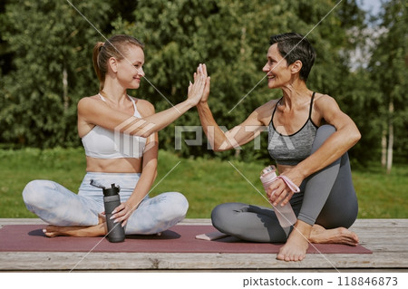 Caucasian blonde daughter and her black haired sporty mom sitting on deep dusty pink yoga mat, holding bottle of pure water and highing five to each other Caucasian blonde daughter and her black haired sporty mom sitting on deep dusty pink yoga mat, holding bottle of pure water and highing five to each other 118846873