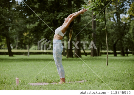 Side view shot of young adult woman standing in Urdhva Hastasana pose on fresh cut lawn in park 118846955