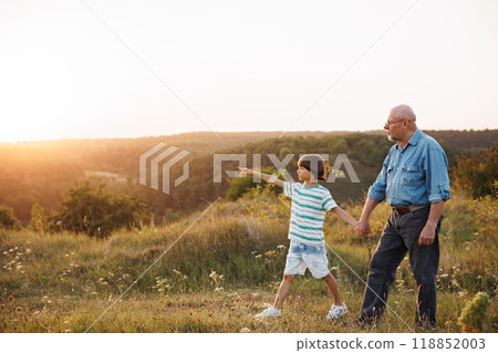 Photo of little boy and his grandfather walking in the field. Boy and grandfather spending time together at summer. Brunette boy wearing striped t-shirt and man blue shirt and glasses. 118852003