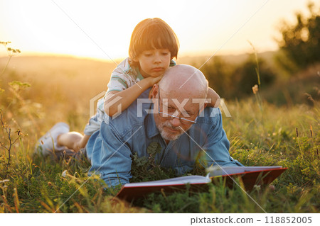 Little boy and his grandfather laying on a grass and reading a book in the field. Boy and grandfather spending time together at summer. Brunette boy wearing striped t-shirt and man blue shirt and Little boy and his grandfather laying on a grass and reading a book in the field. Boy and grandfather spending time together at summer. Brunette boy wearing striped t-shirt and man blue shirt and 118852005