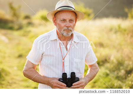 Portrait of a mature man with binoculars in a field. Bearded caucasian man looking through a binoculars in a field at summer. Man wearing striped t-shirt and a hat. 118852010