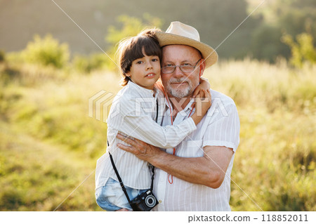 Photo of little boy and his grandfather standing and hugging in the field. Boy and grandfather spending time together at summer. Brunette boy and man wearing striped shirts. Photo of little boy and his grandfather standing and hugging in the field. Boy and grandfather spending time together at summer. Brunette boy and man wearing striped shirts. 118852011