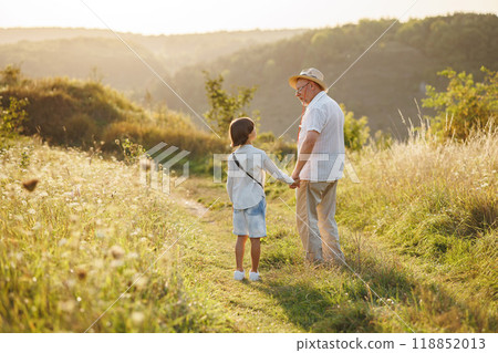 Backside photo of little boy and his grandfather walking in the field. Boy and grandfather spending time together at summer. Brunette boy and man wearing striped shirts. Backside photo of little boy and his grandfather walking in the field. Boy and grandfather spending time together at summer. Brunette boy and man wearing striped shirts. 118852013