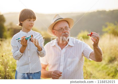 Portrait of little explorer and his grandfather playing with two toys plane in a field. Boy and grandfather spending time together at summer. Brunette boy and man wearing striped shirts. Portrait of little explorer and his grandfather playing with two toys plane in a field. Boy and grandfather spending time together at summer. Brunette boy and man wearing striped shirts. 118852015