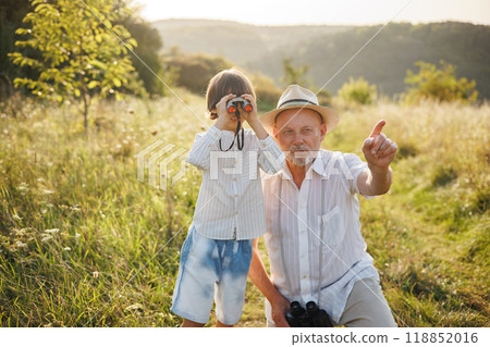 Portrait of little explorer and his grandfather with binoculars in a field. Boy and grandfather spending time together at summer. Brunette boy and man wearing striped shirts. Portrait of little explorer and his grandfather with binoculars in a field. Boy and grandfather spending time together at summer. Brunette boy and man wearing striped shirts. 118852016