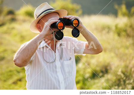 Portrait of a mature man with binoculars in a field. Bearded caucasian man looking through a binoculars in a field at summer. Man wearing striped t-shirt and a hat. 118852017
