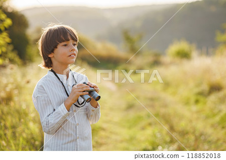 Portrait of little explorer with binoculars in a field. Little caucasian boy looking through a binoculars in a field at summer. Brunette boy wearing striped shirt. 118852018