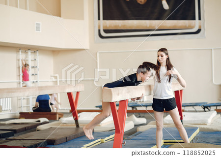 Child gymnastics balance beam. Girl gymnast athlete during an exercise horizontal bar in gymnastics competitions. Coach with child. 118852025